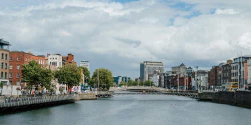 Scenic view of the River Liffey running through Dublin City with a mix of historic and modern buildings on either side, a pedestrian bridge in the centre, and a cloudy sky overhead