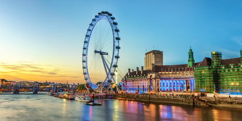 The London Eye at sunset, surrounded by illuminated buildings, as seen from across the Thames.